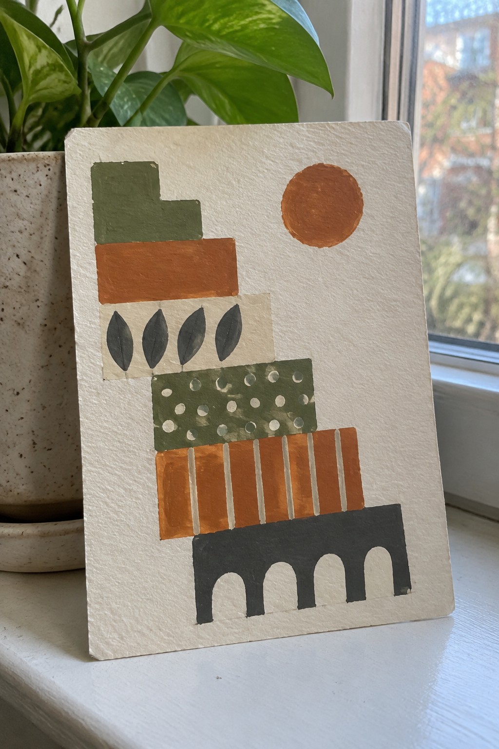 Abstract painting with orange sun, green shapes, leaves, and black arches beside a plant