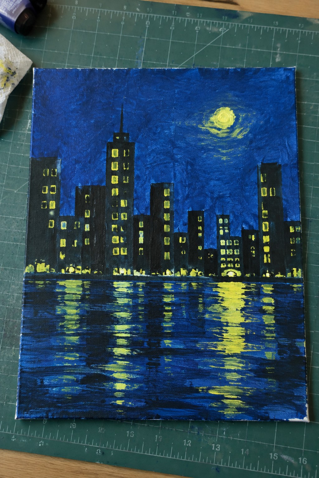 Nighttime cityscape painting with glowing yellow windows reflected in blue water under moon