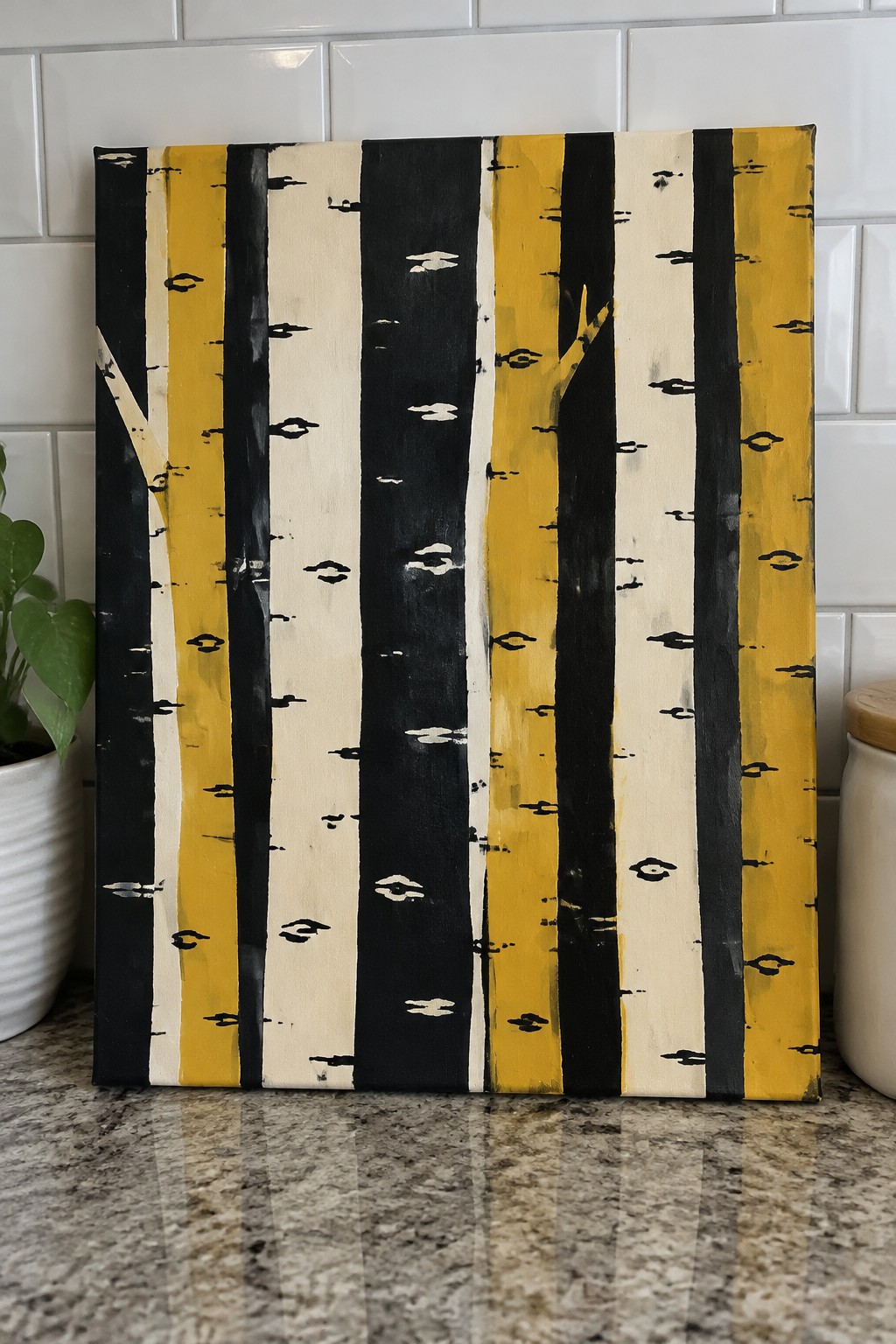 A square canvas painting with vertical stripes of black, yellow, and off-white representing stylized tree trunks, placed on a granite countertop.