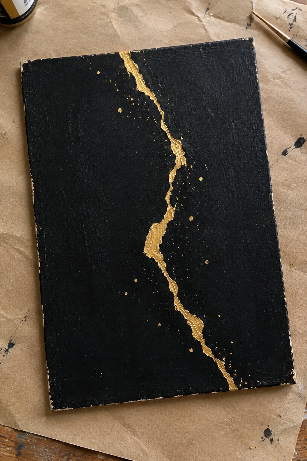 Black canvas with a jagged vertical gold line and scattered gold dots resting on brown paper beside a paint jar and brush.