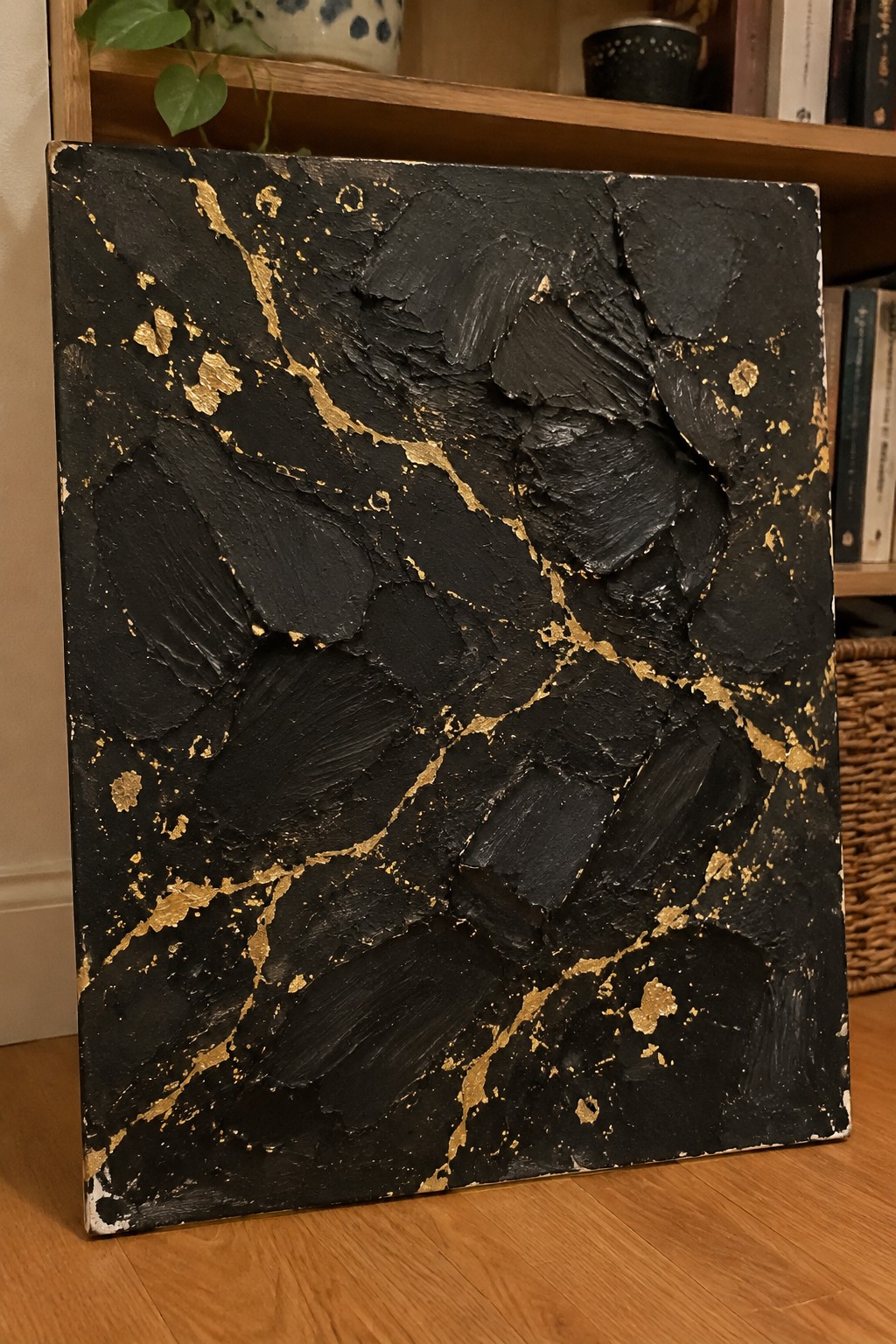 Textured black abstract painting with gold accents on wooden floor before bookshelf