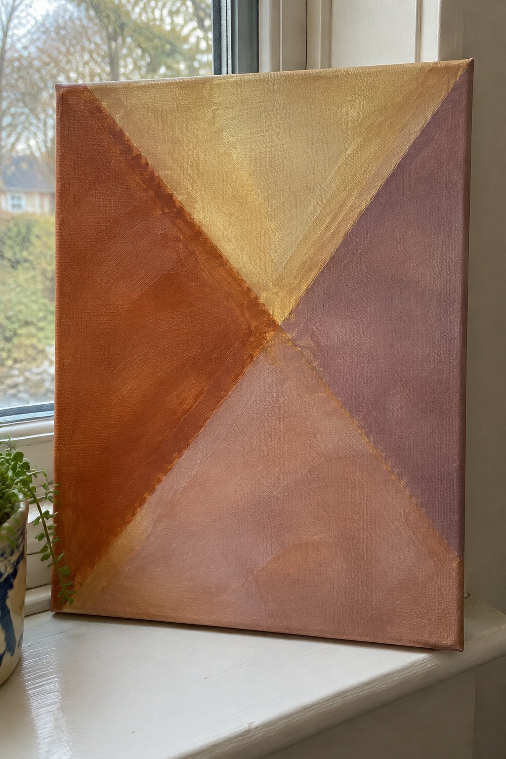 Abstract geometric painting with orange, gold, and mauve triangles on windowsill beside plant