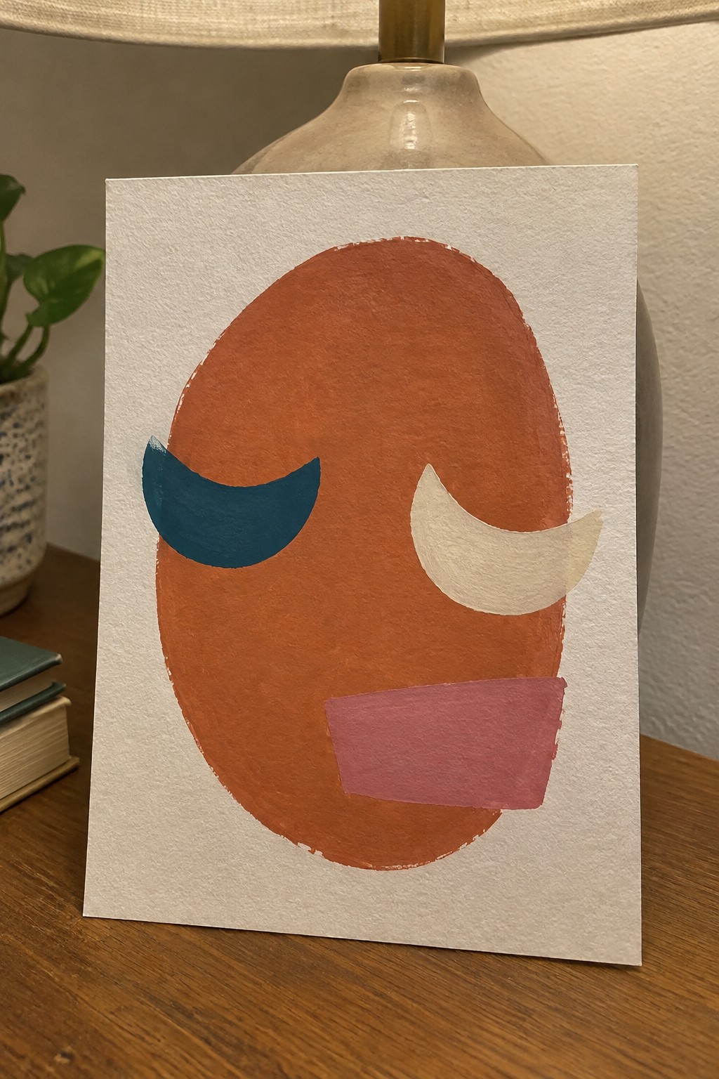 Abstract orange oval face painting with teal eye, white crescent, and pink mouth on paper