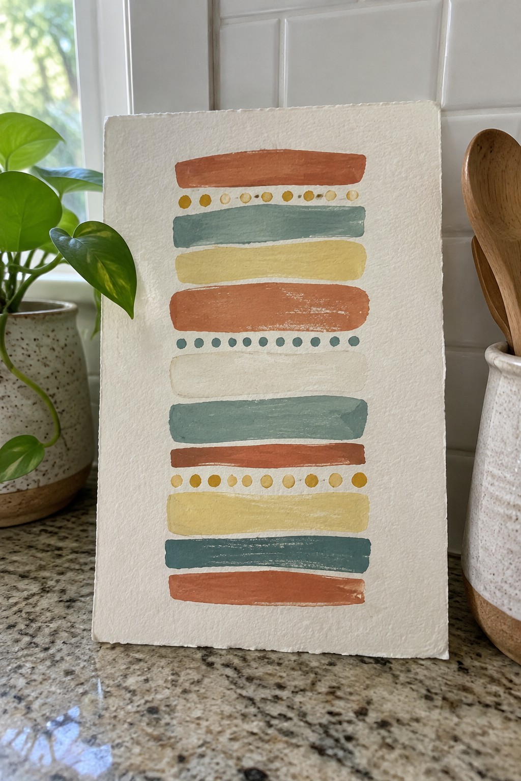 Abstract painting with earth-toned stripes and dots on white paper atop marble counter
