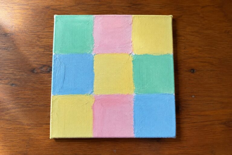 Pastel nine-square grid painting on canvas atop wooden table with plant leaf