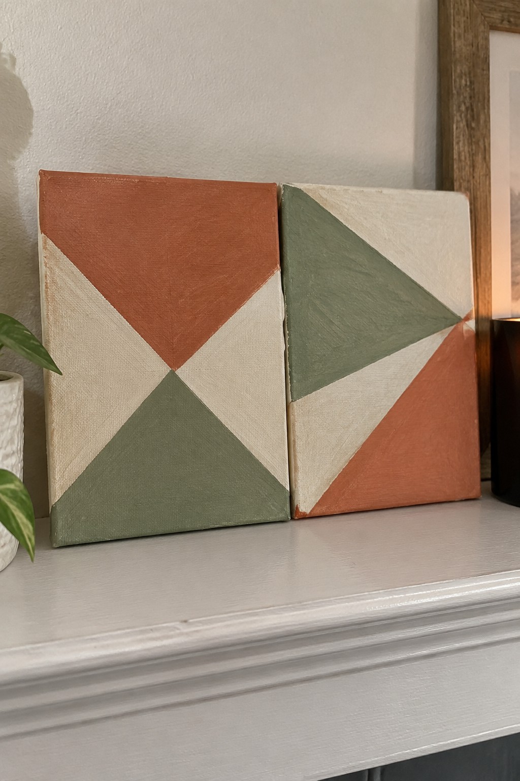 Two geometric canvases with triangular patterns in orange, sage green, and cream on a mantel