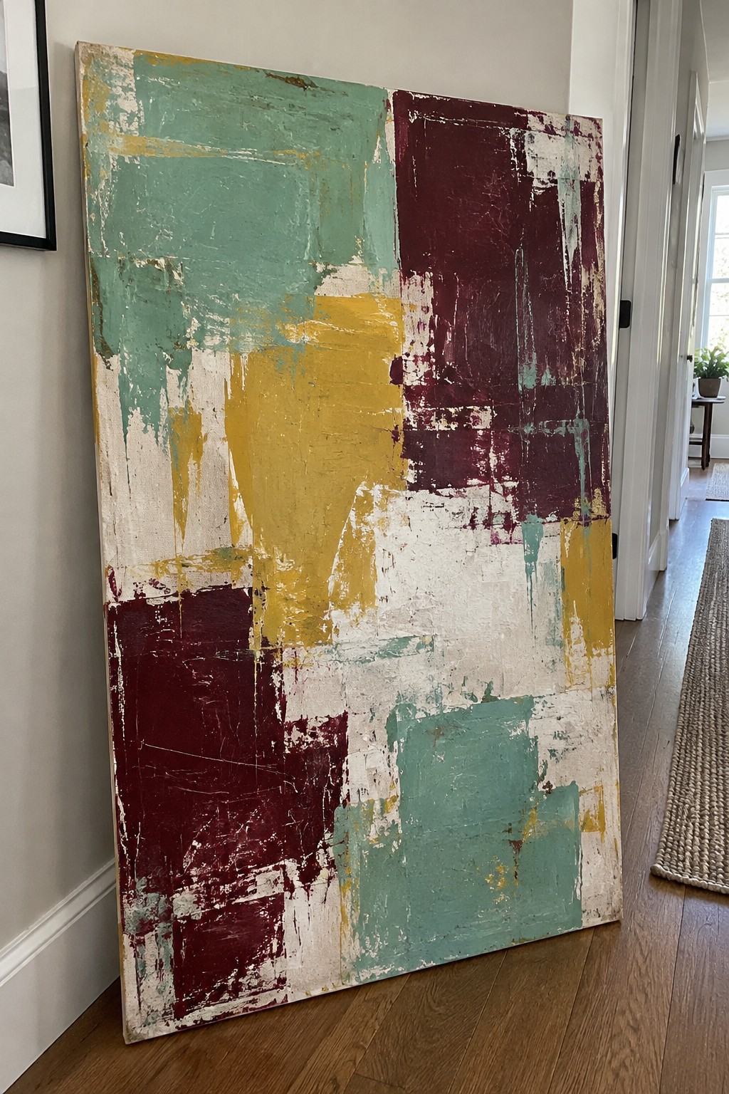 Abstract painting with teal, maroon, yellow and white brushstrokes leaning against hallway wall