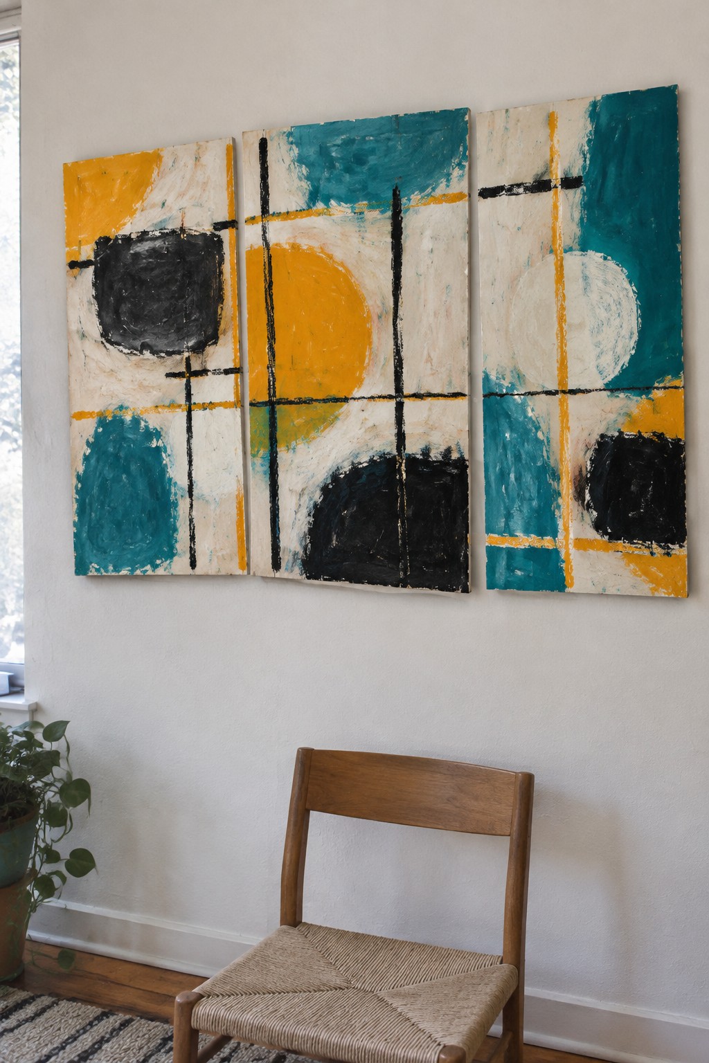 Three abstract paintings in teal yellow black above wooden chair on white wall