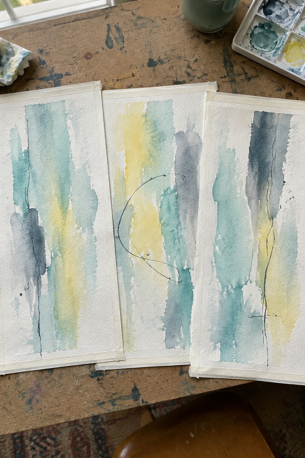 Three abstract watercolors with blue, green, and yellow washes on a wooden table