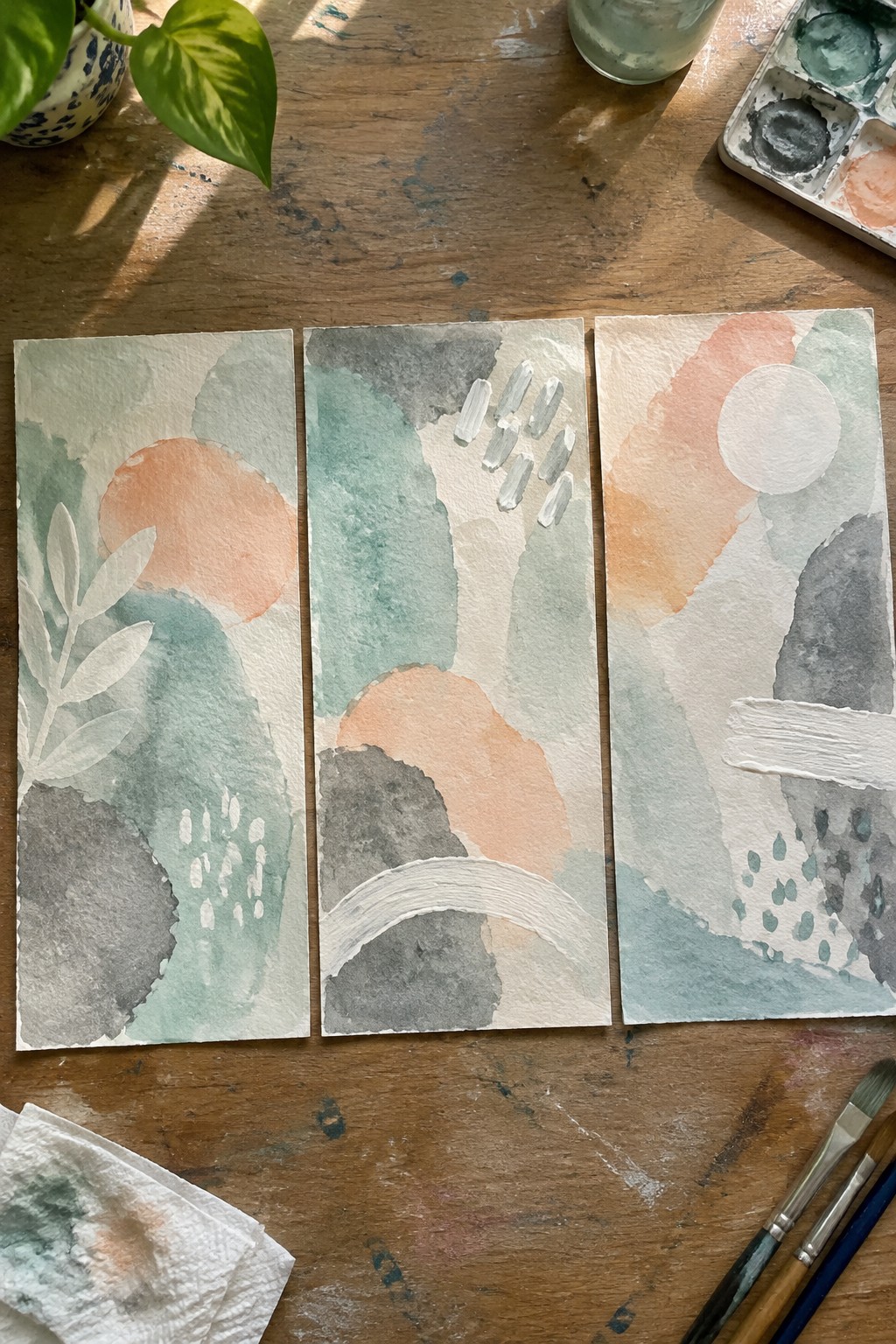 Three abstract watercolors with soft green, peach, and gray shapes on wooden table