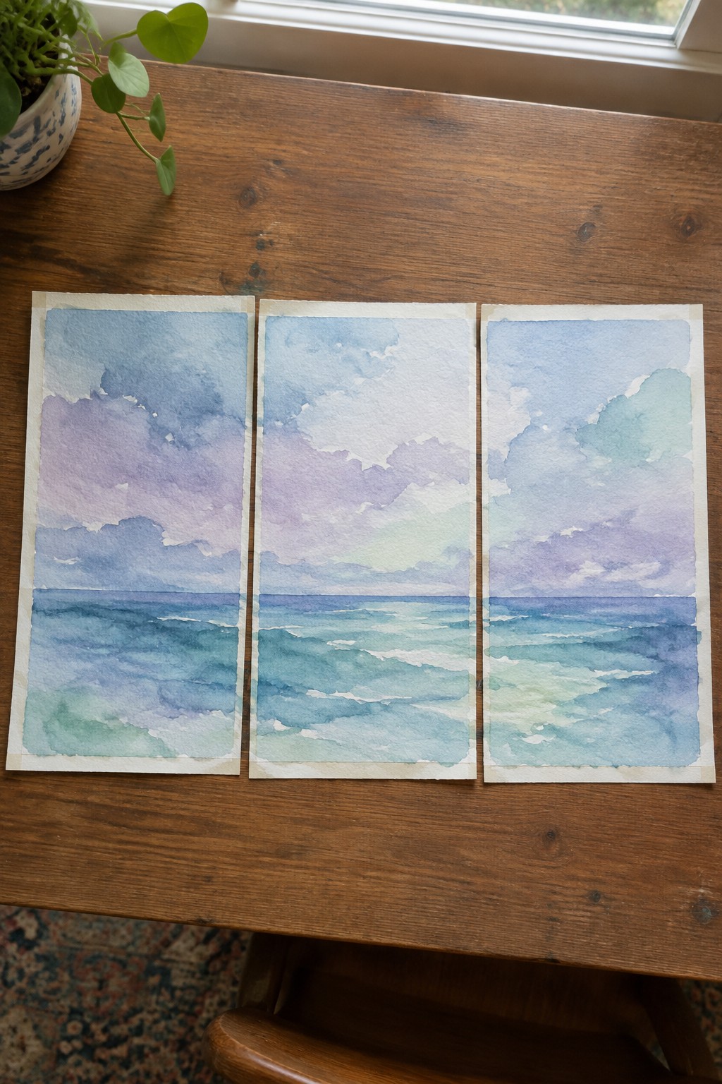 Three watercolor seascapes of ocean waves and skies on a wooden table beside a plant.