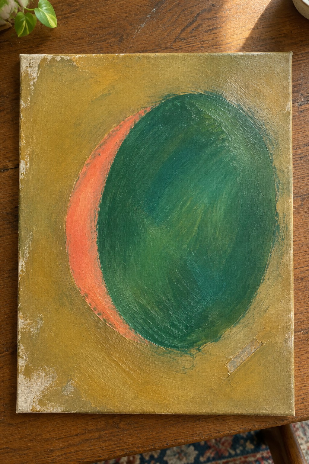 Abstract painting of green oval with red arc on gold background atop wooden table