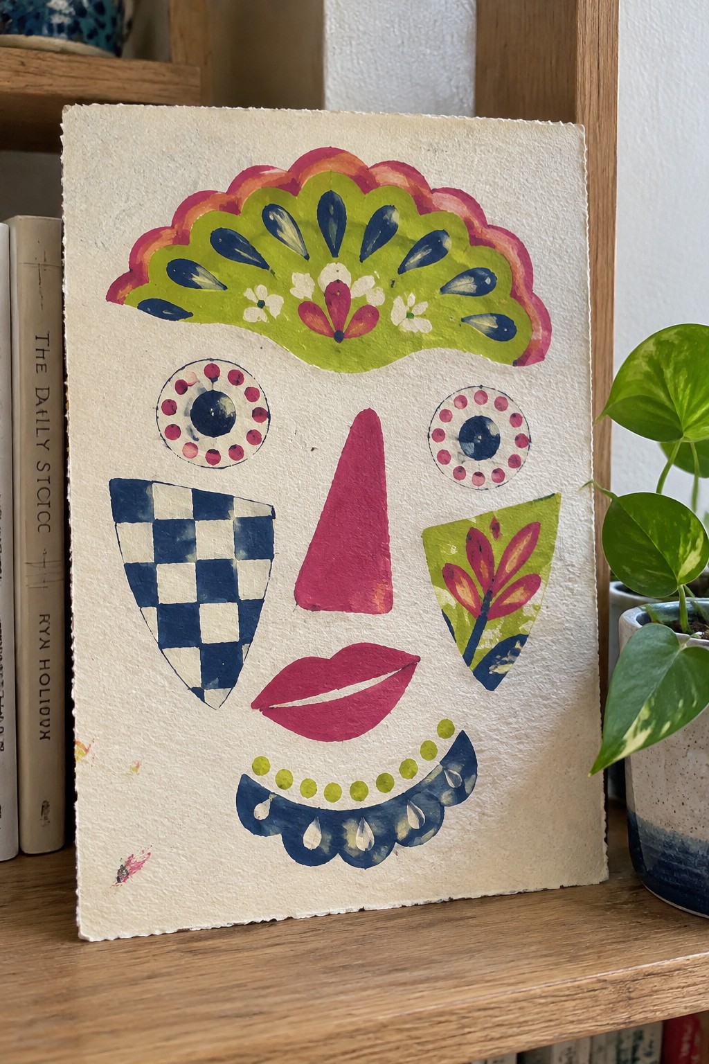 Colorful abstract watercolor face with green headdress, pink nose, and patterned cheeks on shelf.