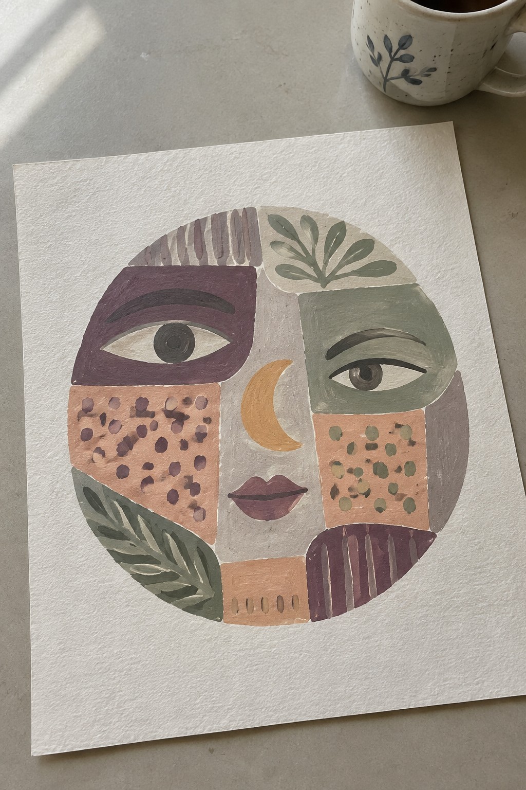 Abstract circular face painting with geometric patterns, leaves, and moon beside speckled mug