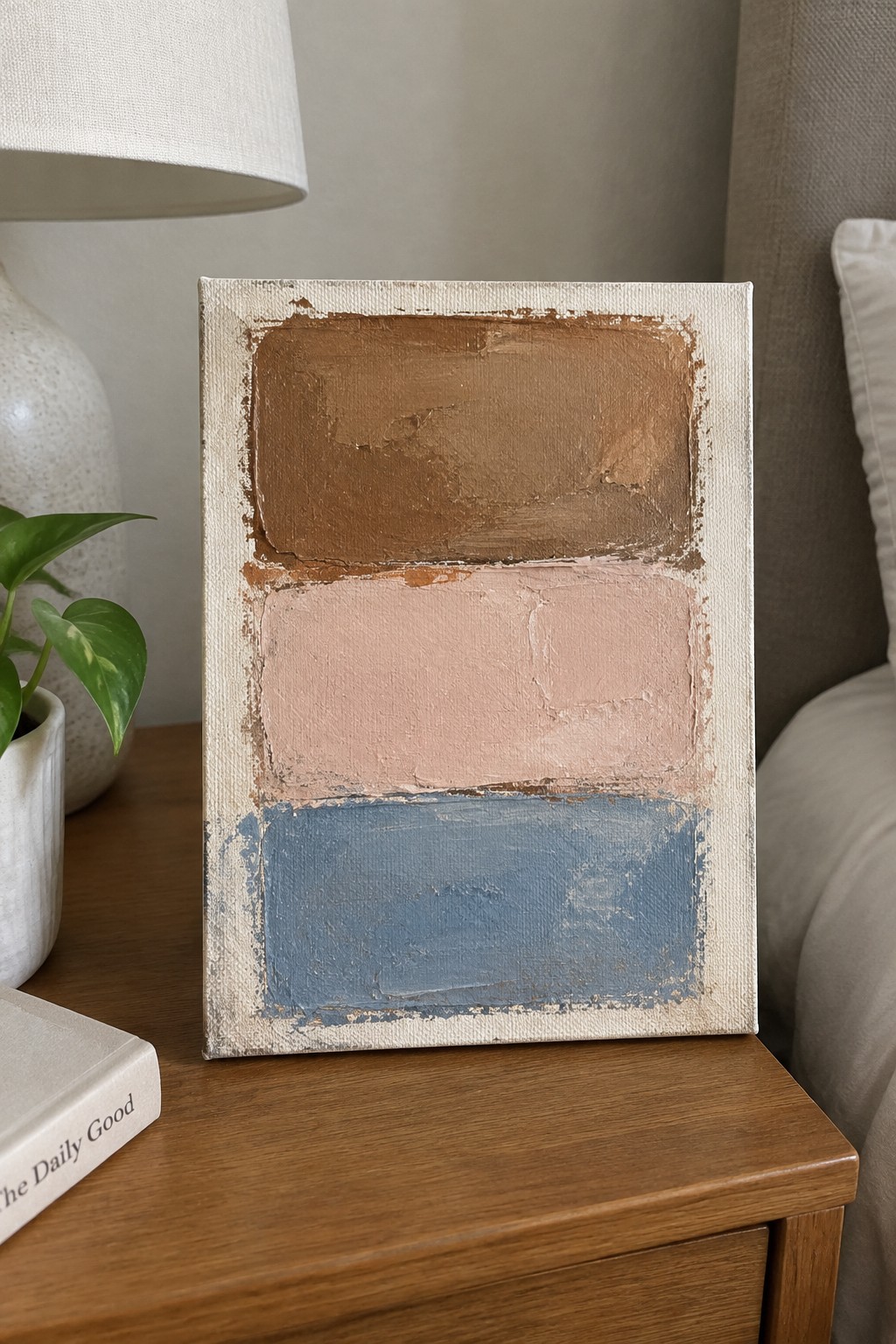 Abstract painting with brown, pink, and blue sections on wooden bedside table beside lamp and plant