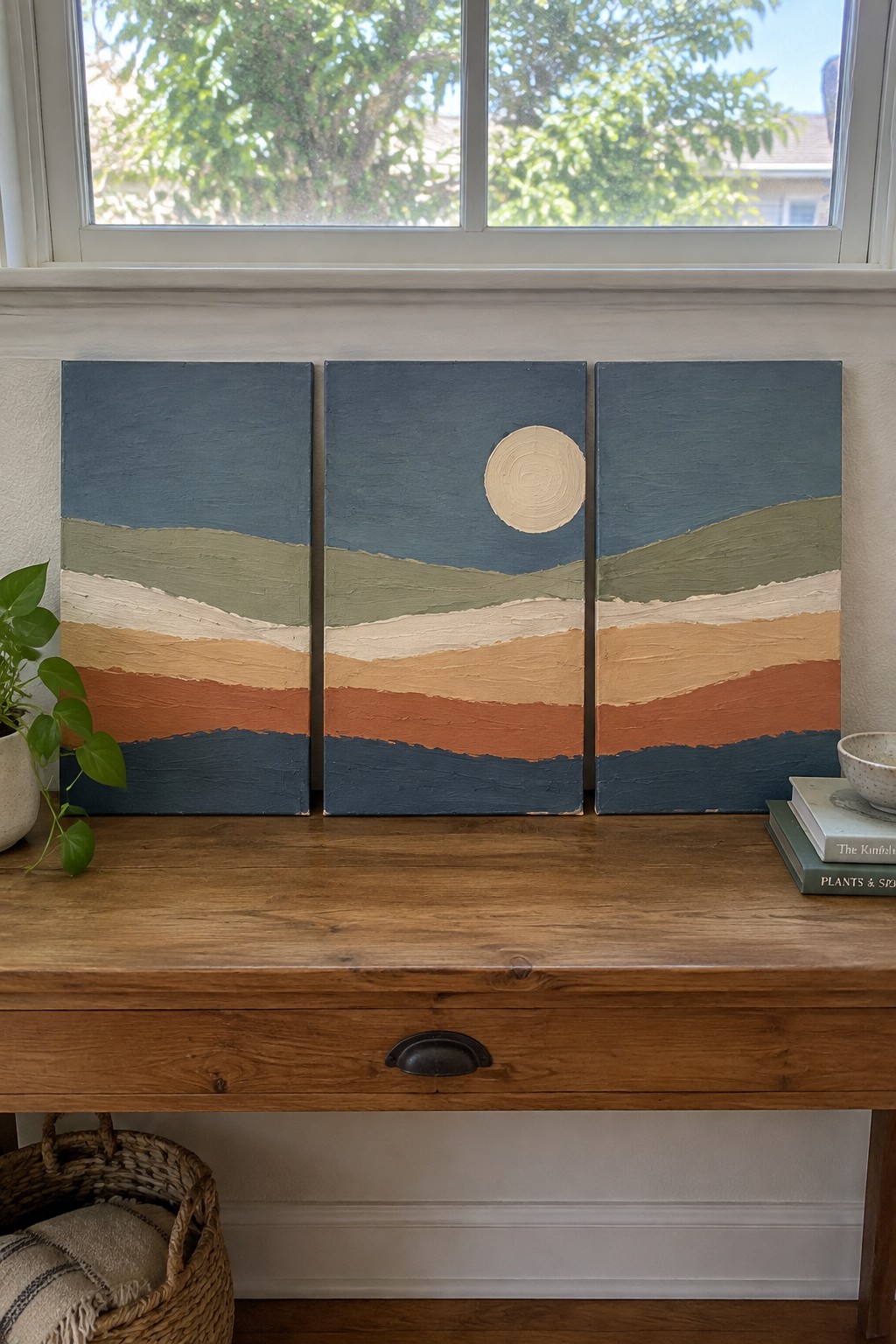 Three abstract landscape paintings on wooden table with plant, books, and basket