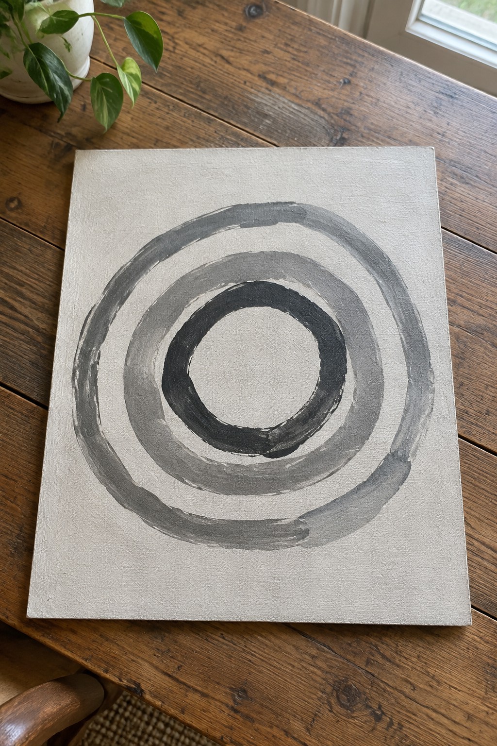 Abstract painting of concentric black and gray circles on canvas atop wooden table with plant