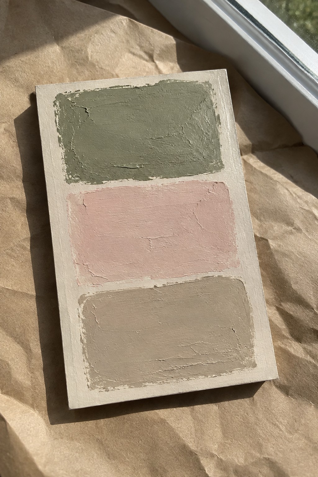 A light wood board displays three horizontal painted sections in muted green, soft pink, and warm gray resting on brown paper.