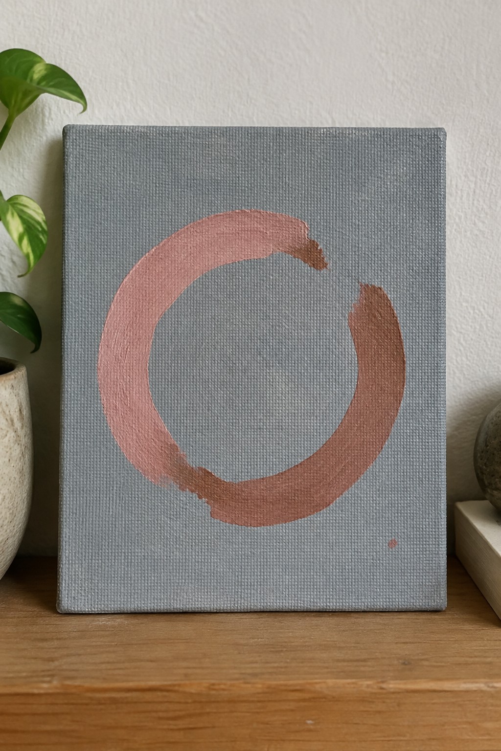 Minimalist acrylic painting showing a large incomplete circle in dusty rose with brushstrokes on a solid gray canvas and a small dot accent near the bottom right.