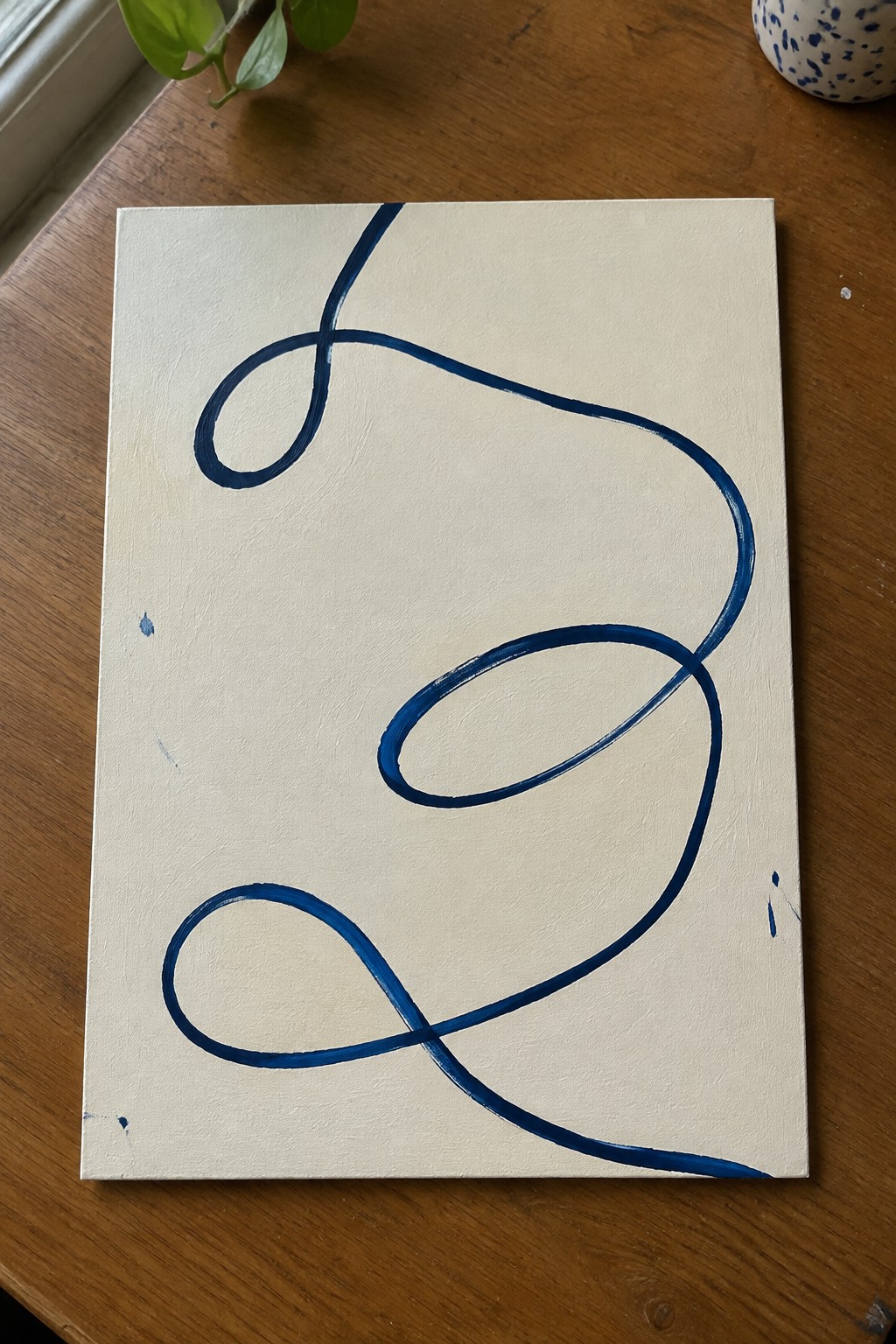 Blue abstract looping lines on off-white canvas atop wooden table with plant nearby