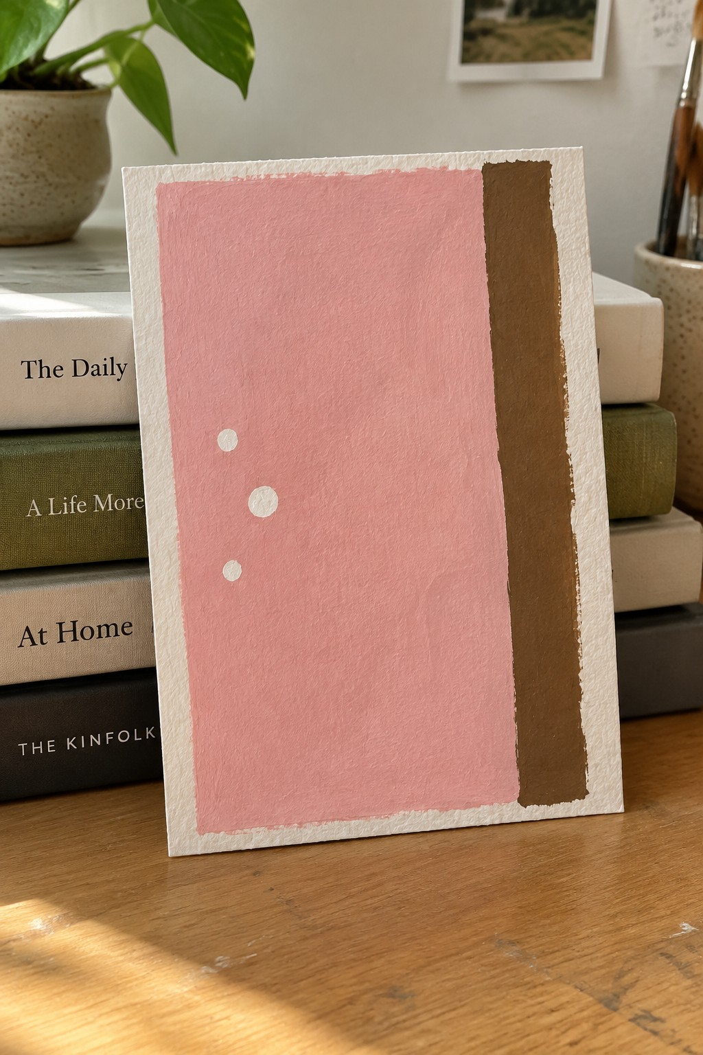 Pink abstract artwork with three white dots and brown stripe against stacked books.