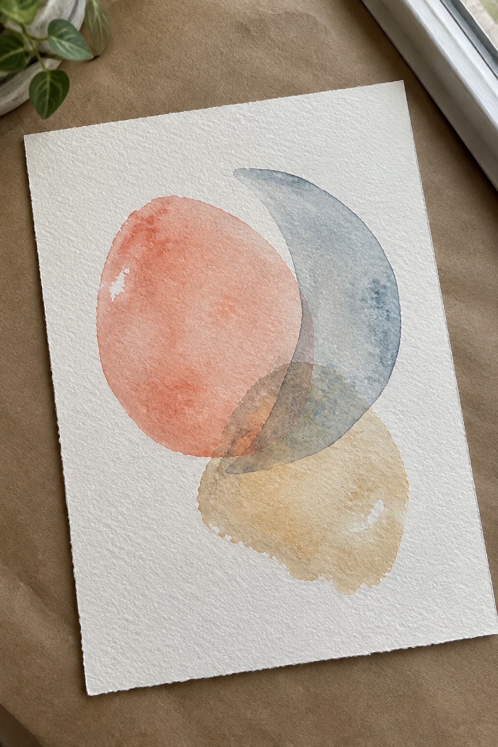 Abstract watercolor with overlapping coral circle, blue crescent, and beige shapes on white paper.