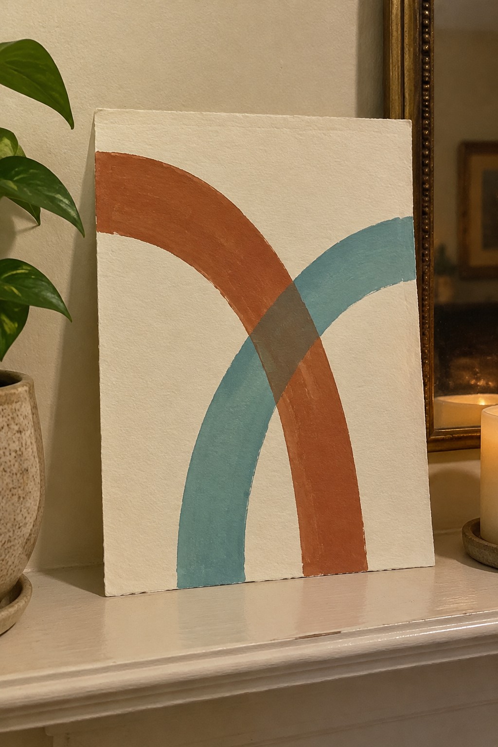 Abstract painting with overlapping terracotta and teal curves on white mantel beside plant and candles