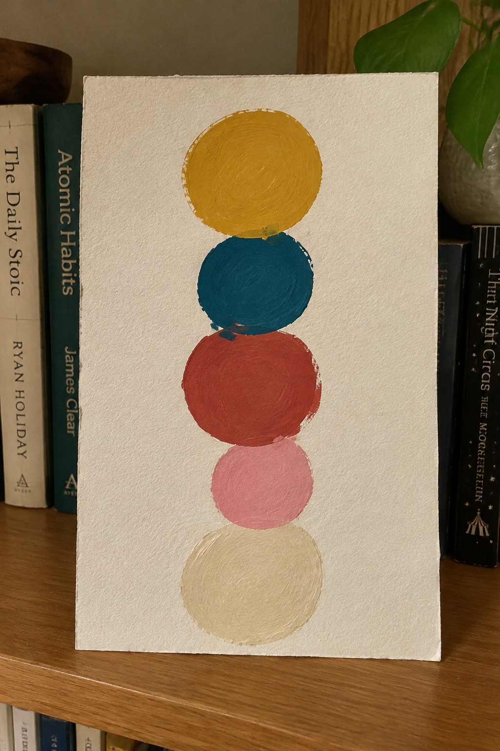 Five stacked circles painted yellow teal red pink beige on bookshelf with books.