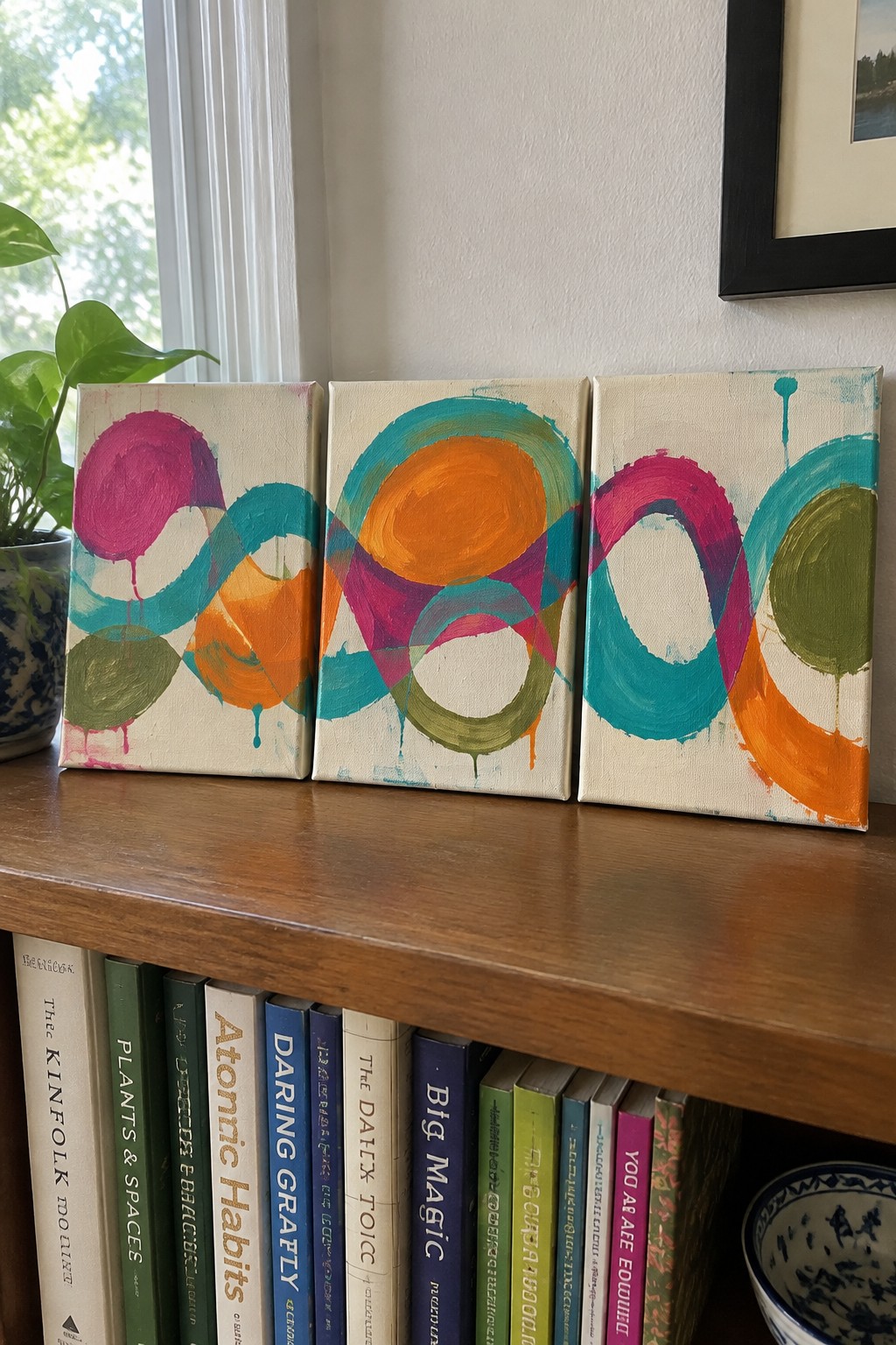 Three abstract paintings with colorful circles and curves on a wooden shelf above books