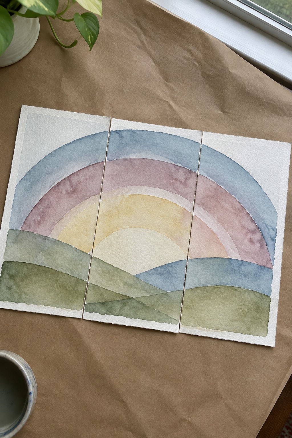 Pastel watercolor rainbow triptych over layered green hills on brown paper