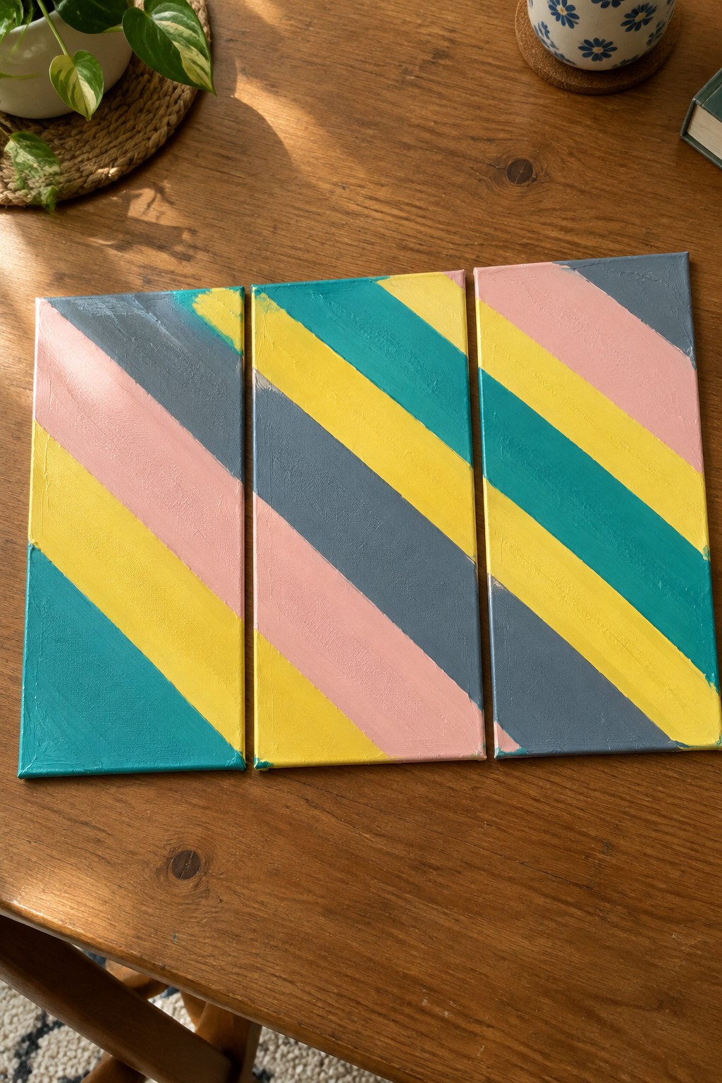 Three abstract canvases with pink, yellow, teal, and gray diagonal stripes on a table