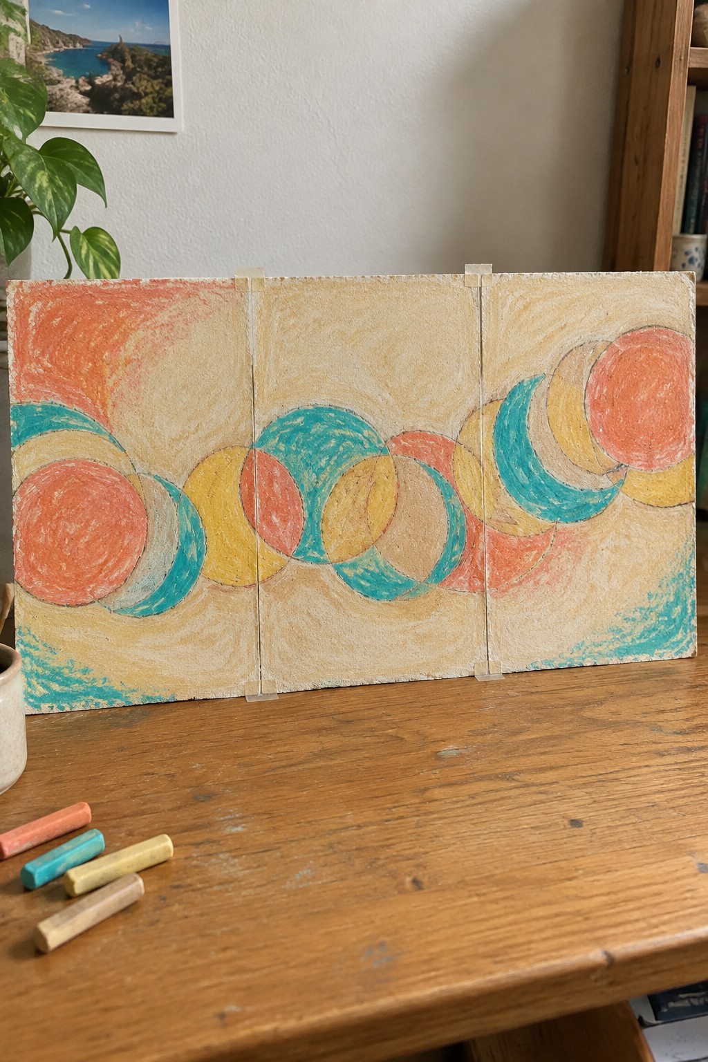 Three-panel abstract artwork with overlapping colorful circles on wooden table beside pastels