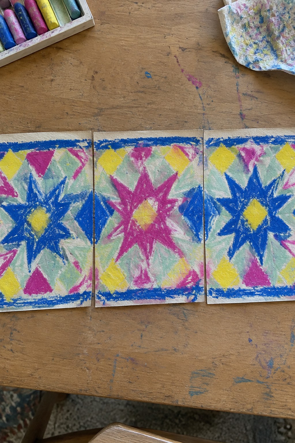 Three side-by-side rectangular panels on a wooden table show matching geometric designs with large blue and pink stars on a field of yellow, pink, and green diamonds, all framed by thick blue borders.