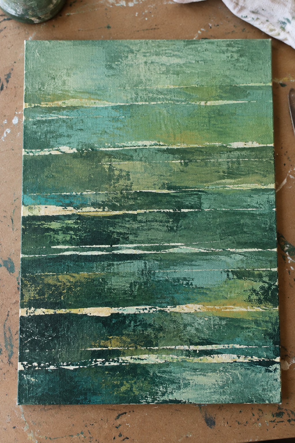 Abstract canvas with horizontal green teal and gold brushstrokes on paint-splattered surface