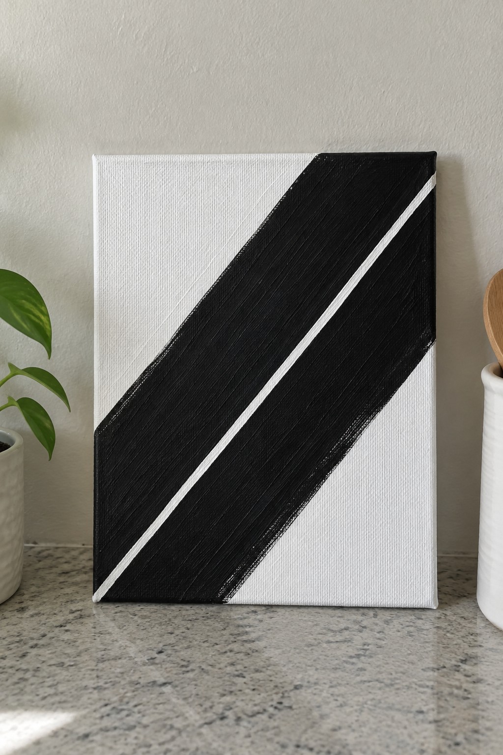 Abstract black-and-white diagonal stripe painting on canvas atop speckled countertop with plant nearby