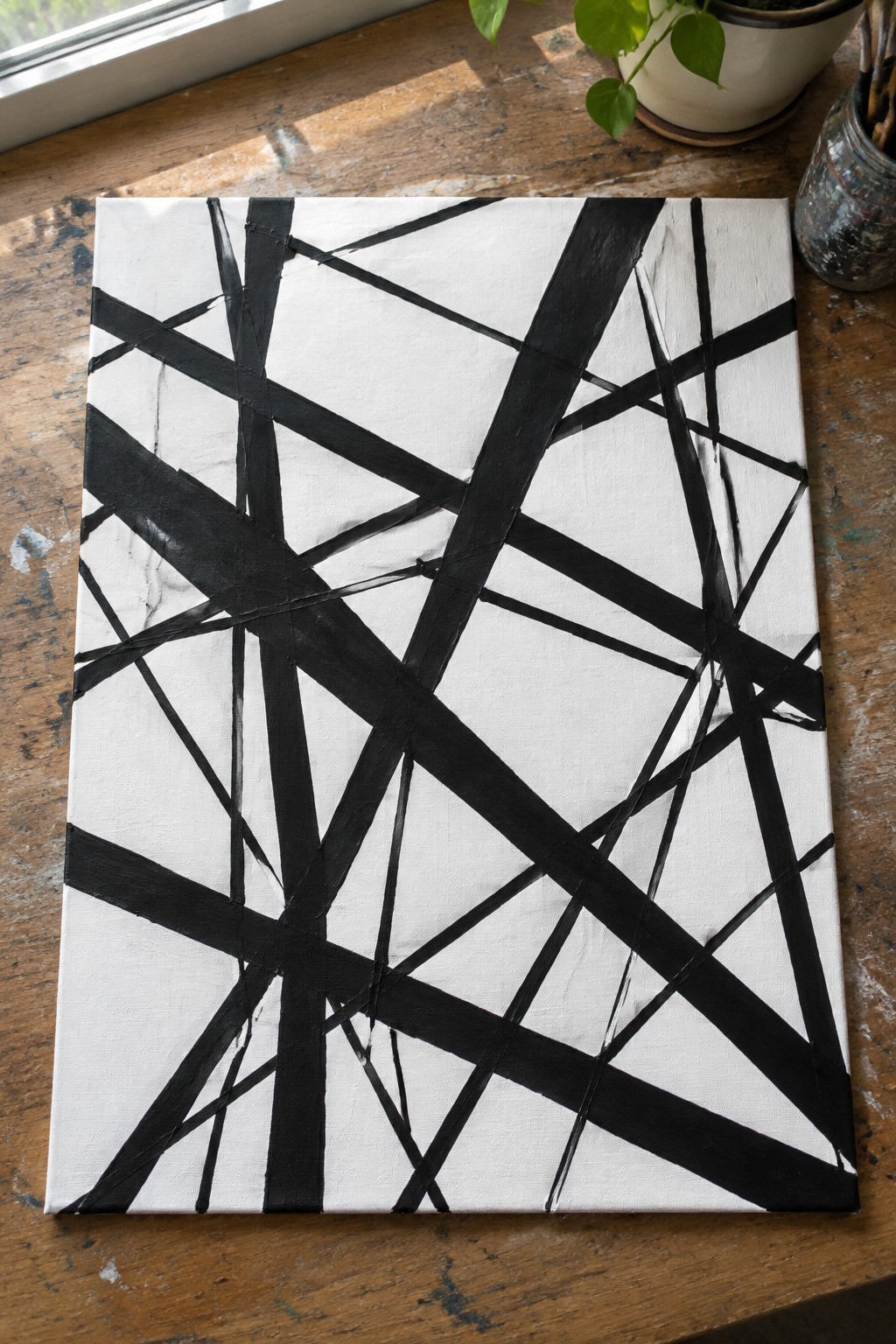 Abstract black lines crisscross white square canvas on wooden table with potted plant.
