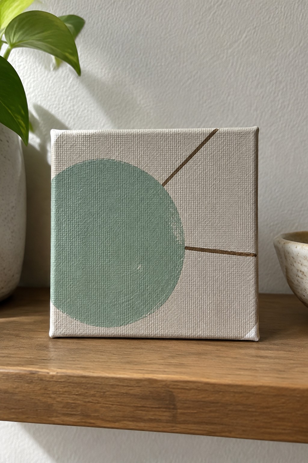 Abstract painting with green circle and brown lines on wooden shelf beside plant