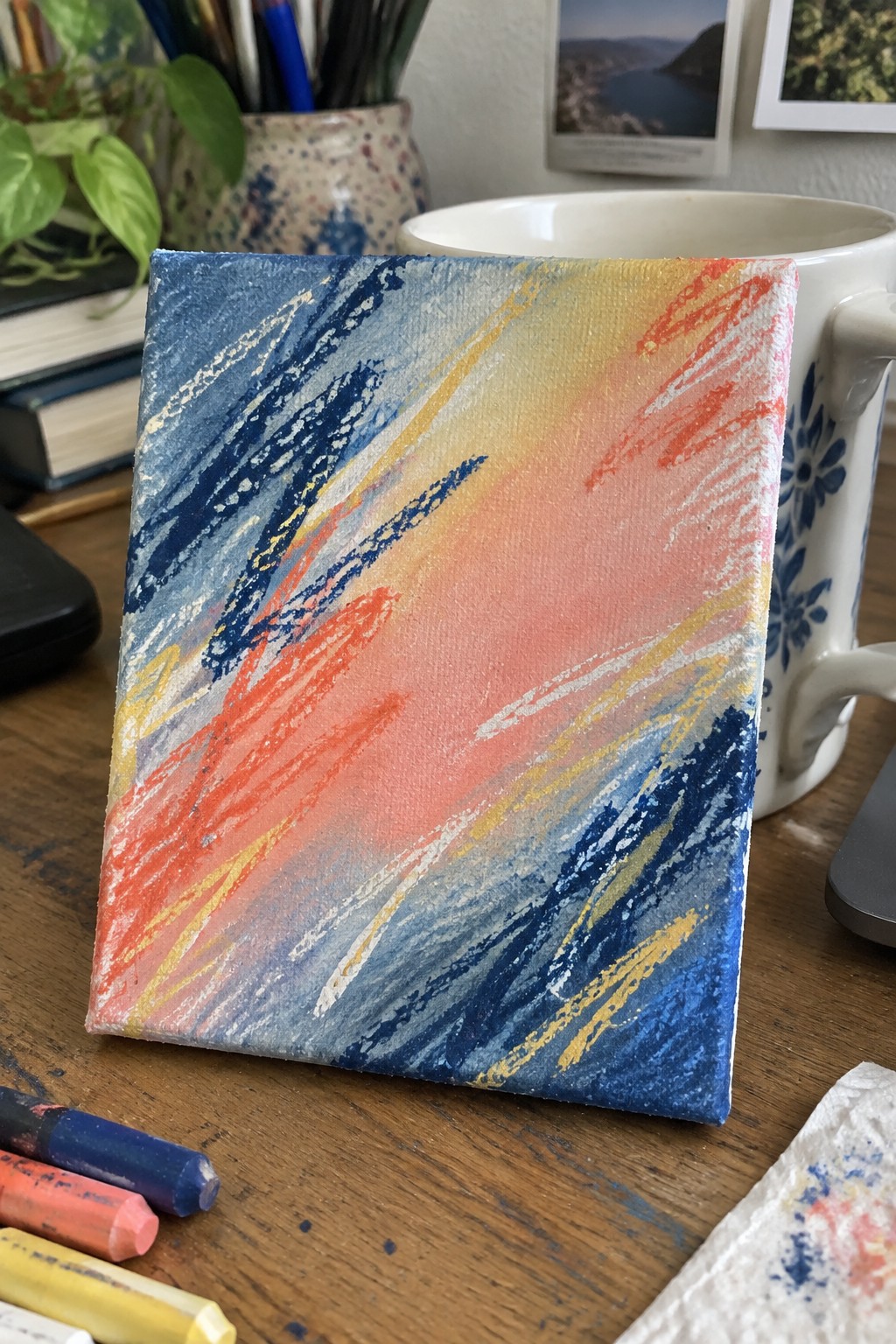 Abstract painting on small canvas against floral mug with oil pastels on wood table