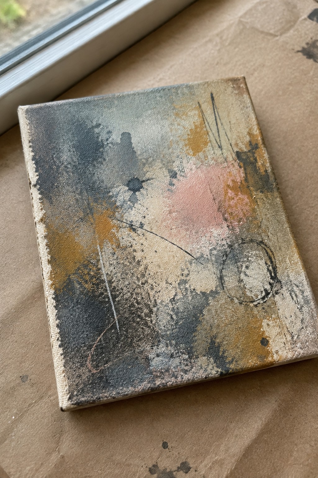 Abstract canvas with gray, orange, pink splatters and marks on brown paper by window