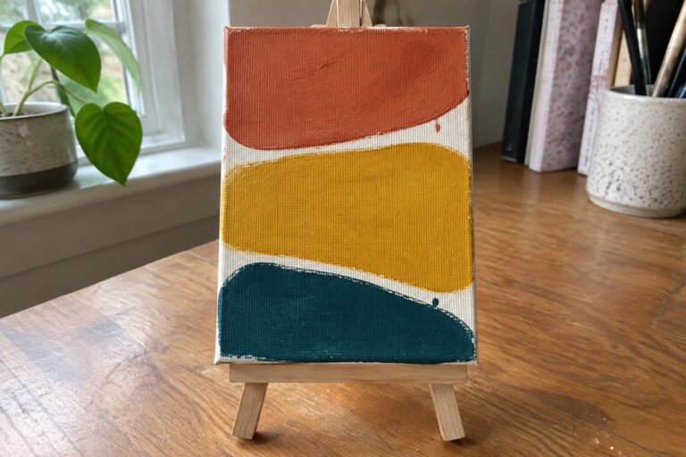Abstract acrylic painting on mini canvas with dripping orange, yellow, teal shapes on easel