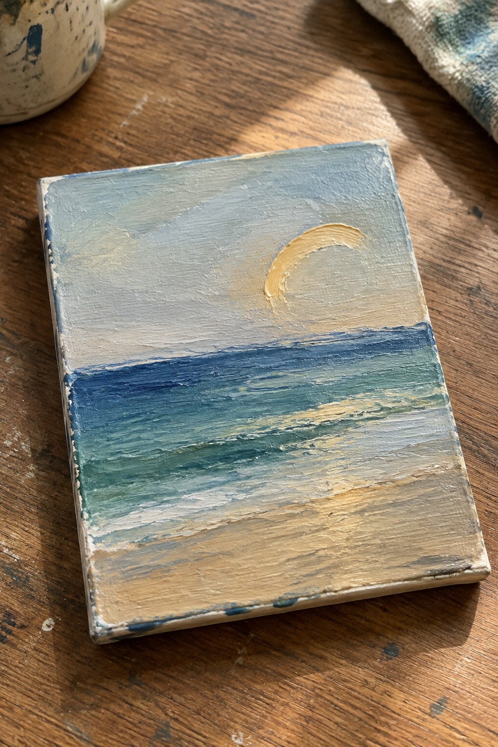 Small seascape painting of ocean waves and crescent moon on wooden table beside mug