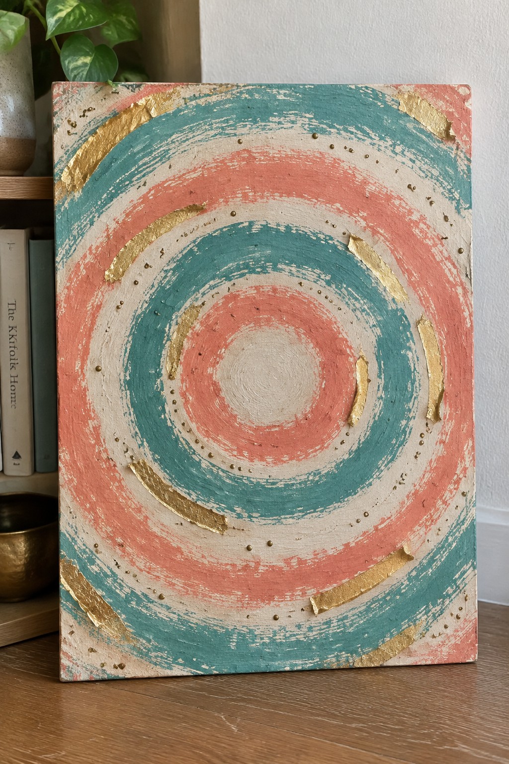 Abstract painting with teal, coral, and beige concentric circles accented by gold leaf on wood