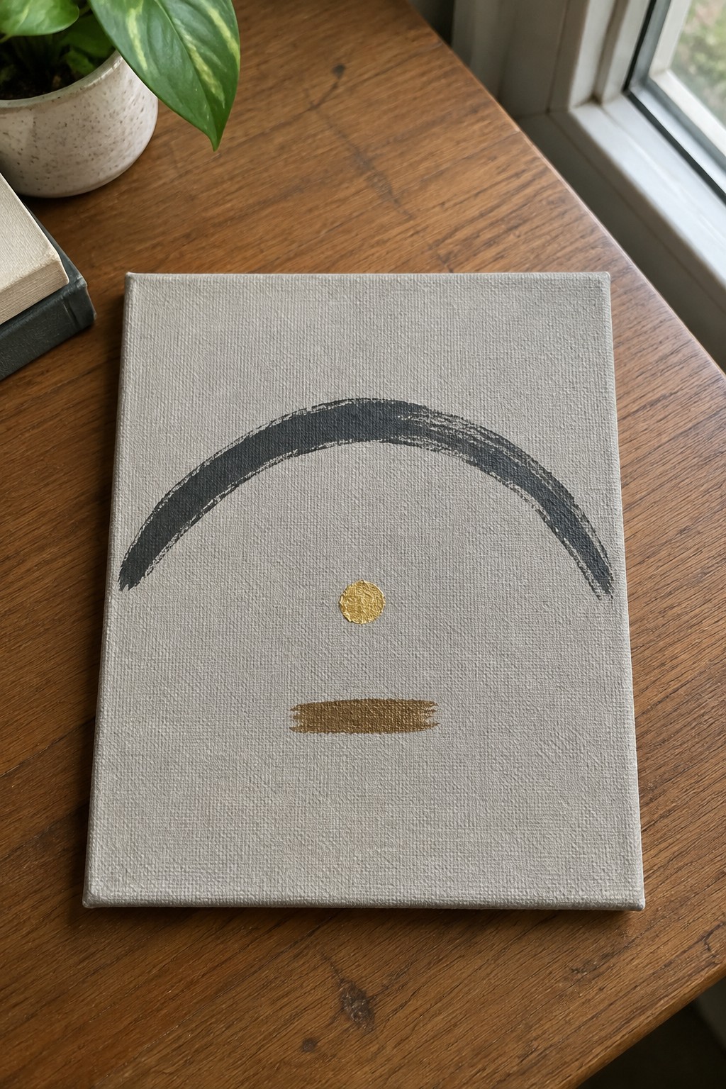 Minimalist canvas with gray arch, gold dot, and line on wooden table beside plant
