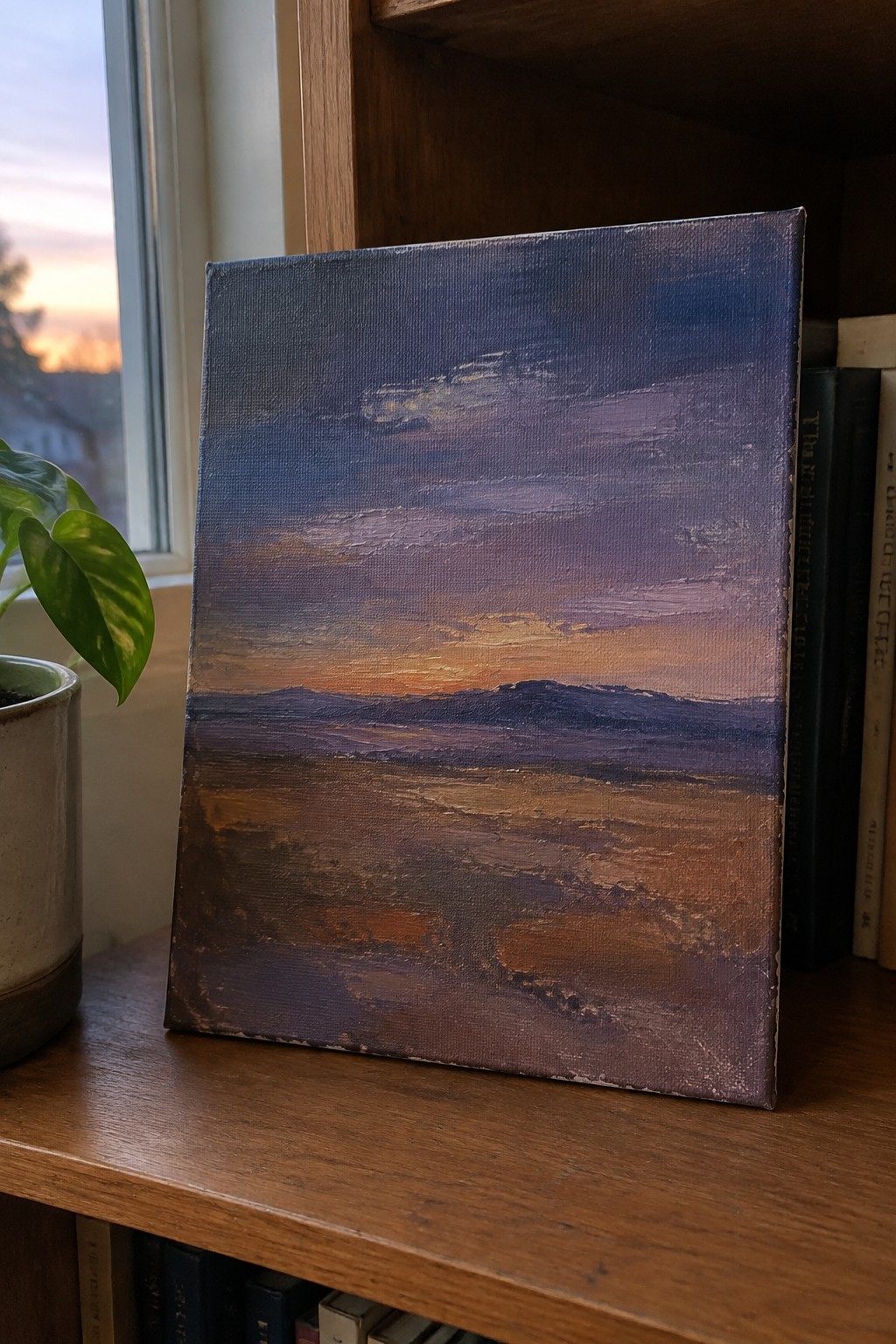 Purple and orange sunset landscape painting on wooden shelf beside potted plant and books.