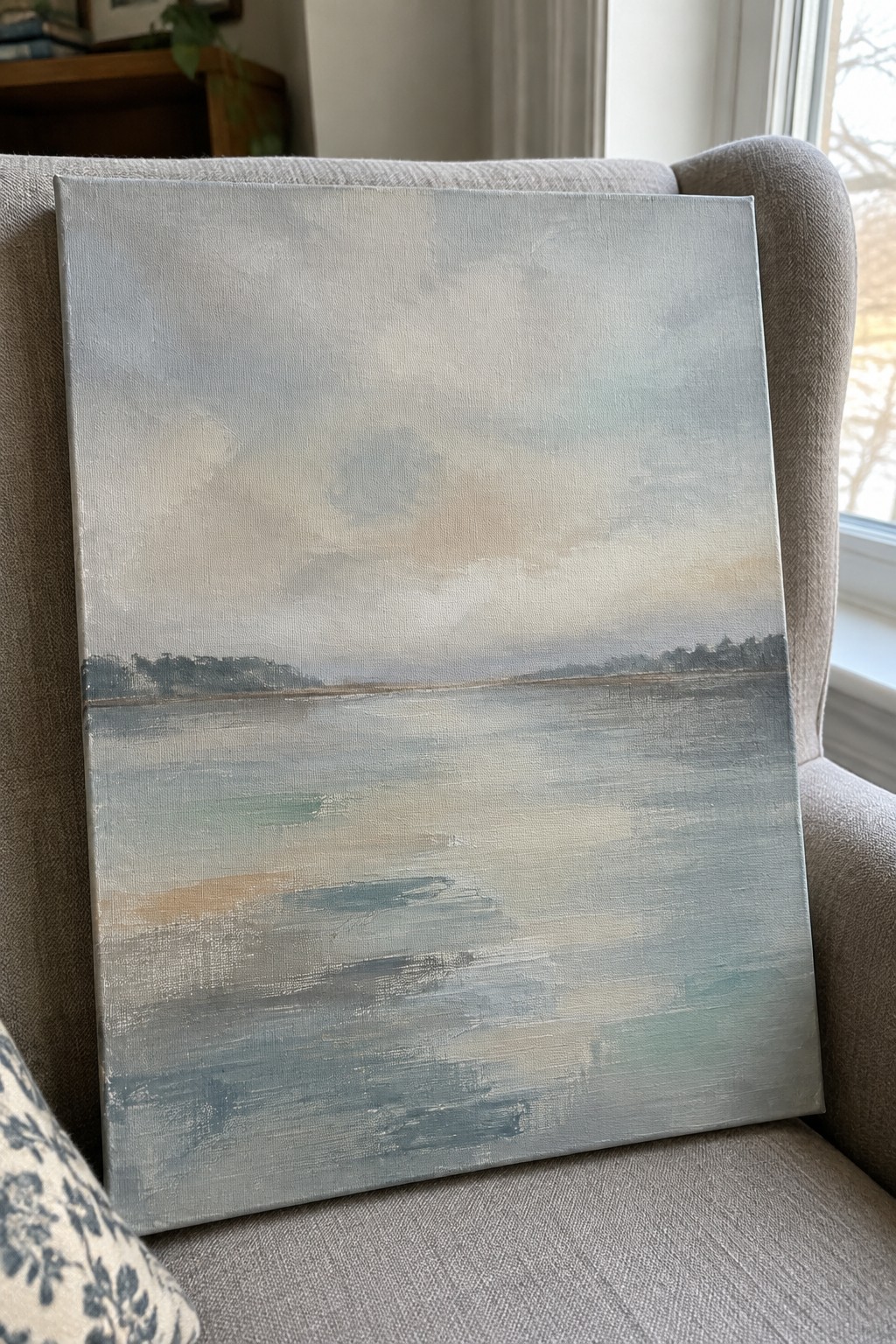 Abstract blue-gray seascape painting resting on gray upholstered armchair near window