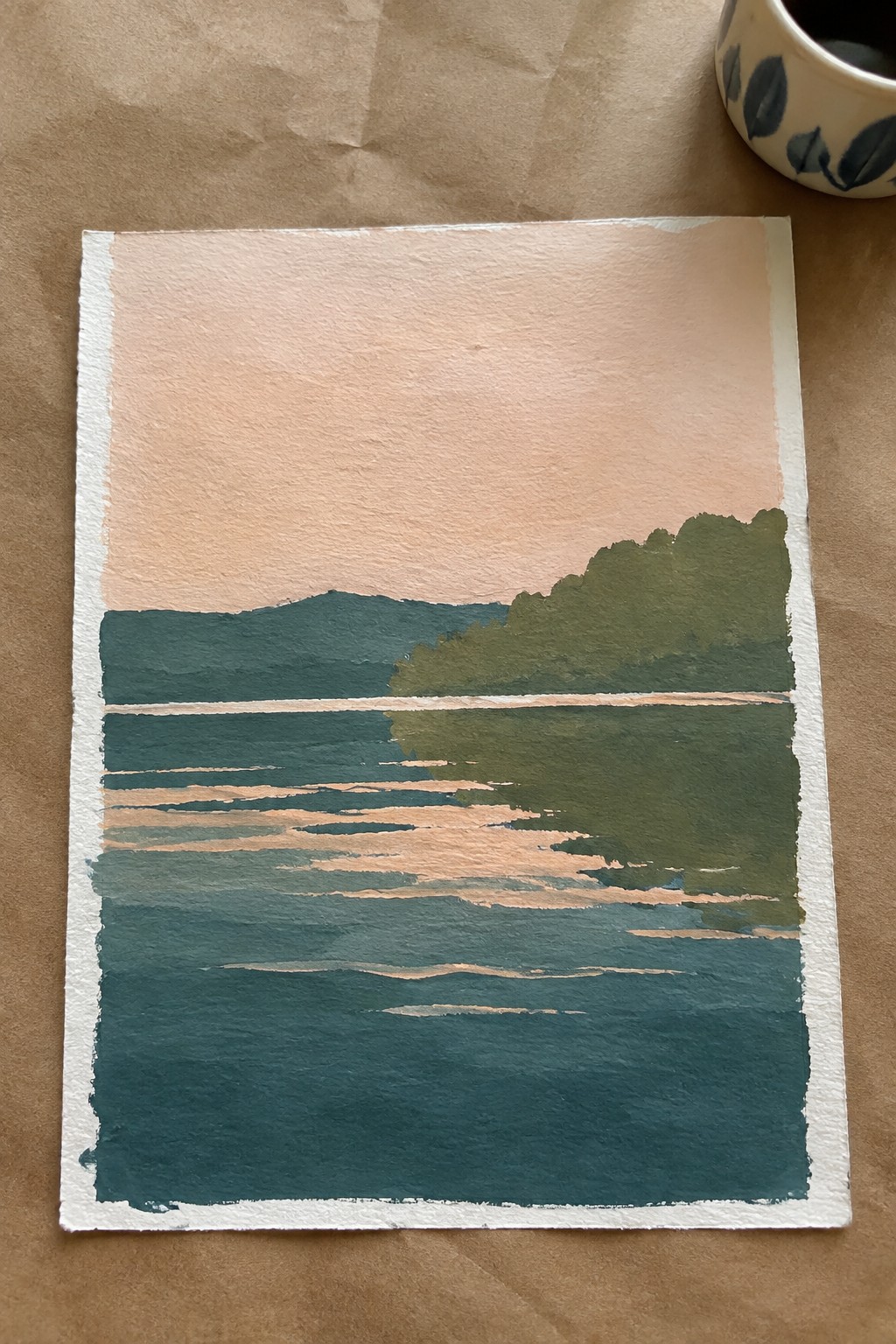 Watercolor landscape with peach sky, green hills, and striped teal water beside patterned cup on paper.