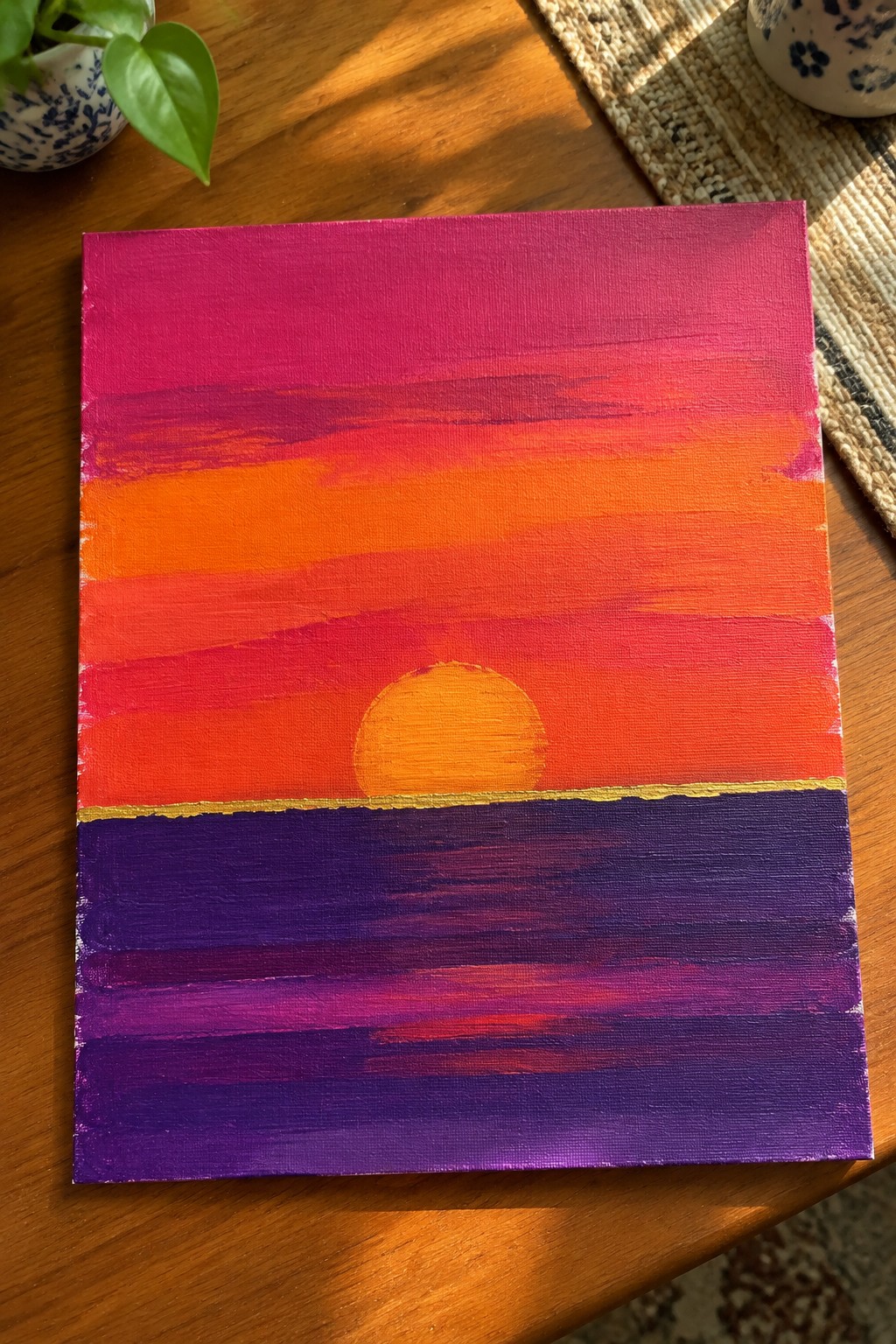 Vibrant sunset painting with orange sun over gold horizon above purple water on wood table