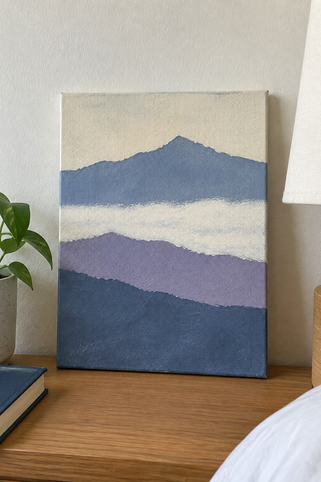 Abstract blue-purple mountain painting on canvas beside plant and blue book on wood.