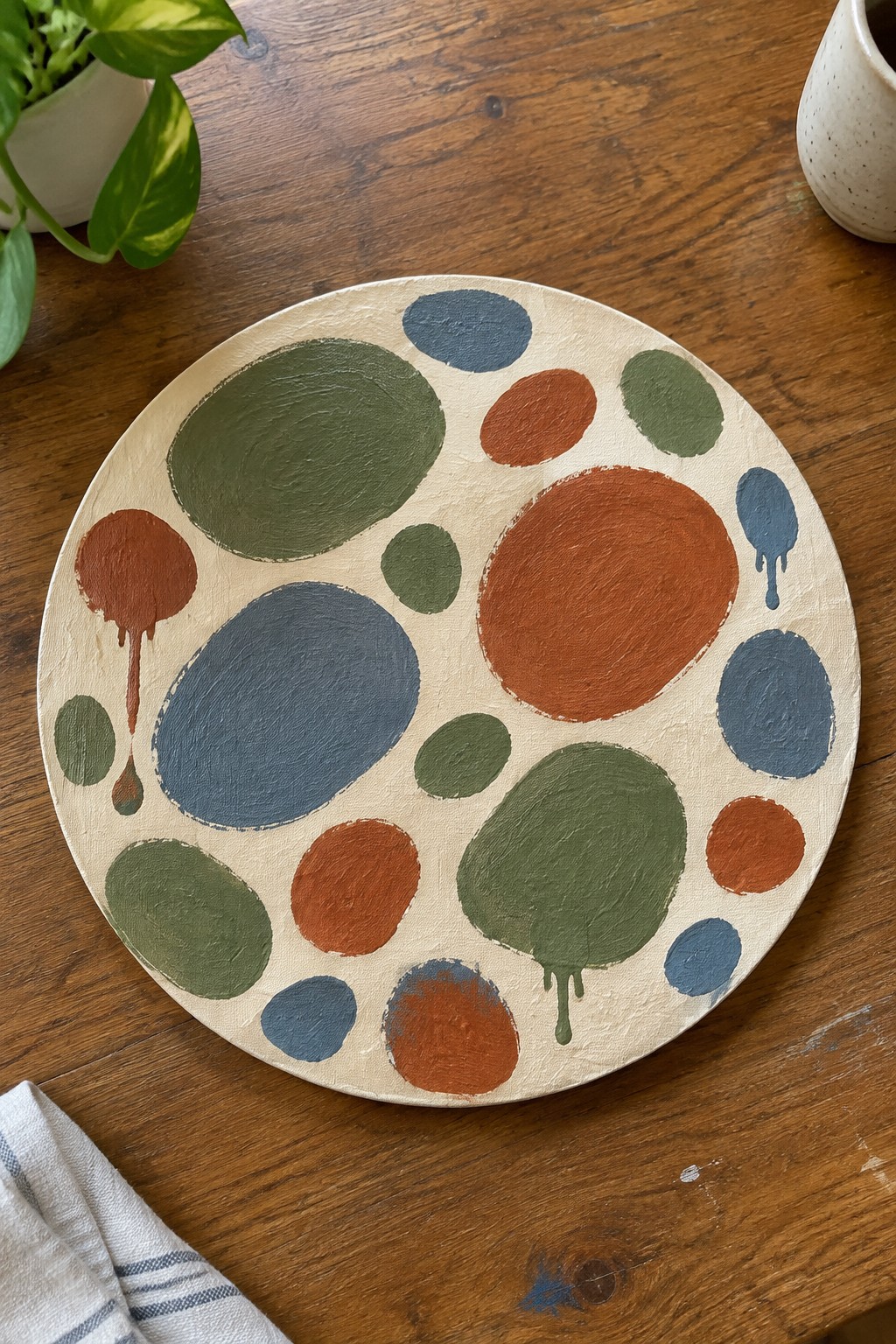 Round canvas with abstract blue, green, and orange circles and drips on wood table