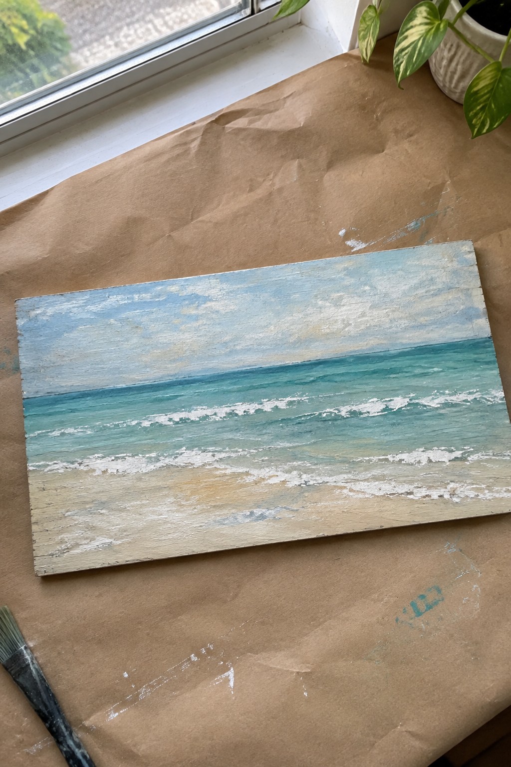 Seascape painting of turquoise waves on wooden panel beside paintbrush on paper